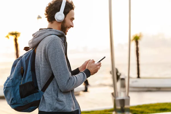 Uśmiechnięty Młodzieniec Słuchawkach Korzystający Telefonu Komórkowego Podczas Spaceru Promenadzie — Zdjęcie stockowe