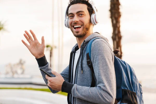 Smiling guy in headphones waving hand and using mobile phone while walking on promenade