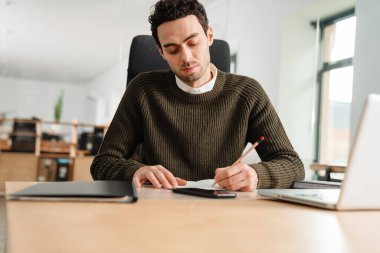 Bir ofiste dizüstü bilgisayar ve belgelerle çalışan girişimci notlar alıyor.