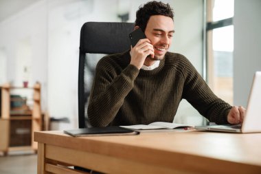 Ofiste dizüstü bilgisayar ve belgelerle çalışan, cep telefonuyla konuşan gülümseyen girişimci.