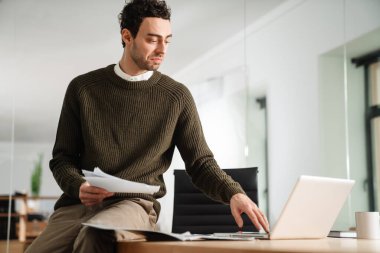Ofiste çalışan kendine güvenen genç yönetici, elinde belgeler, dizüstü bilgisayar kullanıyor.