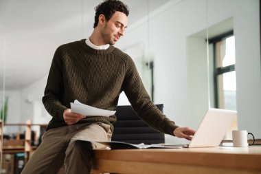 Ofiste çalışan kendine güvenen genç yönetici, elinde belgeler, dizüstü bilgisayar kullanıyor.