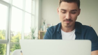 Memnun genç bir adam sanat stüdyosundaki masada oturan dizüstü bilgisayarını kullanıyor.
