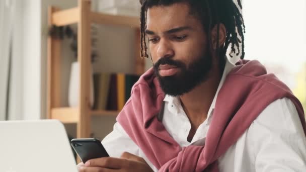 一位有魅力的非洲裔美国人坐在办公室里 一边看着笔记本电脑 一边用他的智能手机 — 图库视频影像