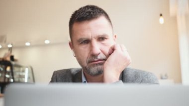 Kablosuz kulaklıklı, ciddi bir yetişkin adam bir kafedeki masada dizüstü bilgisayarını kullanıyor.