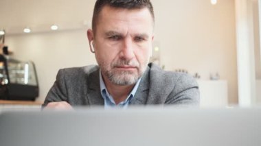 Kablosuz kulaklıklı, odaklanmış yetişkin bir adam bir kafedeki masada oturan dizüstü bilgisayarını kullanıyor.