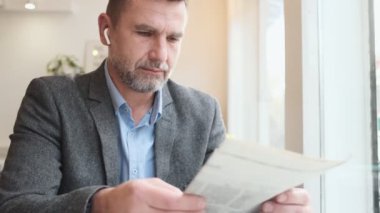 Kablosuz kulaklıklı yakışıklı yetişkin bir adam kafede oturmuş dergi okurken kahve içiyor.