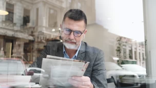 Hombre Adulto Concentrado Con Auriculares Inalámbricos Está Leyendo Revista Sentada — Vídeo de stock
