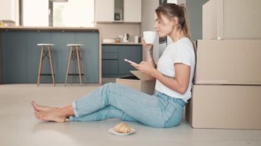 Pozitif bir kadın, evdeki kutuların yanında otururken akıllı telefonunu kullanıyor ve kahve içiyor.