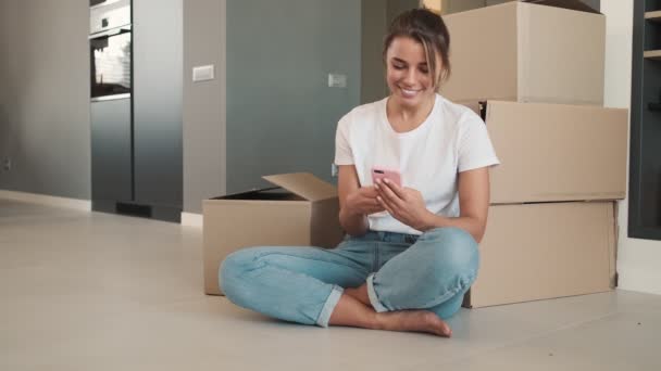 Усміхнена Жінка Використовує Свій Смартфон Сидячи Підлозі Біля Багатьох Коробок — стокове відео