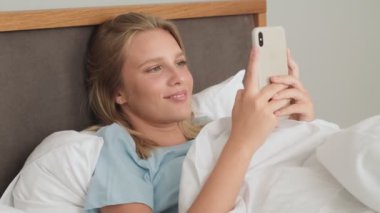 Güzel bir kadın sabah yatağında yatarken akıllı telefonunu kullanıyor.
