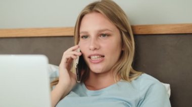 Pozitif bir kadın yatağında dizüstü bilgisayarını kullanırken telefonla konuşuyor.