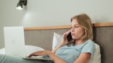 Sakin bir kadın yatağında dizüstü bilgisayarını kullanırken telefonla konuşuyor.