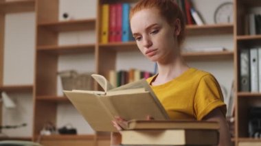 Yoğunlaşmış genç bir kızıl saçlı kadın ofiste masada oturmuş kitap okuyor.