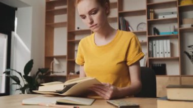 Yoğunlaşmış genç, kızıl saçlı bir kadın ofiste oturmuş kitap okuyor.