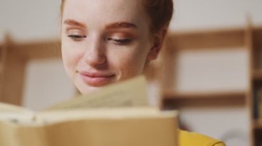 Ofiste masada otururken gülümseyen kızıl saçlı bir kadının yakın çekim görüntüsü kitap okuyor.