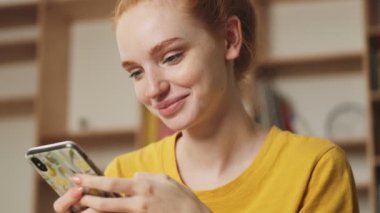 Pozitif, genç, kızıl saçlı bir kadın ofiste çalışırken akıllı telefonunu kullanıyor.