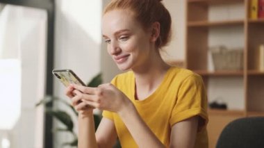 Rüya gören genç bir kızıl saçlı kadın ofiste masada otururken akıllı telefonunu kullanıyor.