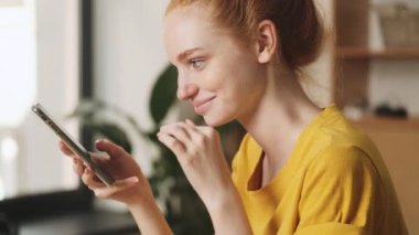 Memnun bir kızıl saçlı kadının yan görüntüsü ofiste masada otururken akıllı telefonunu kullanıyor.