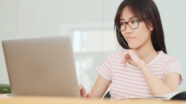 Çekici bir Asyalı kadın, ofiste dizüstü bilgisayarla çalışırken masada oturan defterine bir şeyler yazıyor.