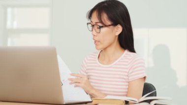 Ciddi bir Asyalı kadın ofiste video bağlantısı ve dizüstü bilgisayar kullanarak grafikler gösteriyor ve konuşuyor.