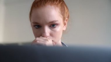 Ciddi kızıl saçlı bir kadın dairesinin içinde dizüstü bilgisayarını kullanıyor.