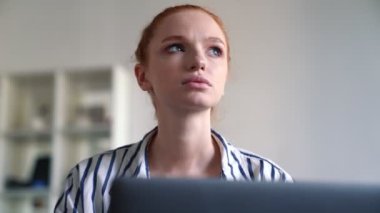 Düşünen kızıl saçlı bir kadın dizüstü bilgisayarını kullanıyor. Dairenin içinde oturuyor.