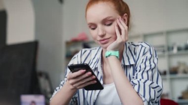 Mutlu, kızıl saçlı bir kadın apartmanda otururken akıllı telefonunu kullanıyor.