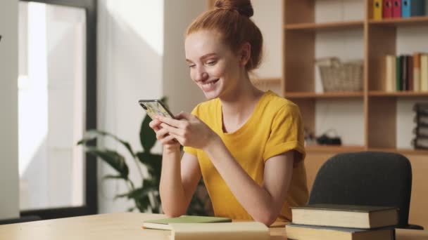 オフィスのテーブルに座っている間 喜んで若い赤毛の女性は彼女のスマートフォンを使用しています — ストック動画