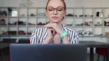 Gözlüklü kızıl saçlı güzel bir kadın dizüstü bilgisayarını kullanıyor. Evin içinde oturuyor.