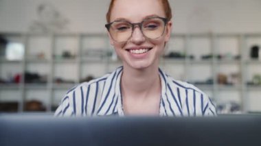 Gözlüklü, gülümseyen kızıl saçlı bir kadın laptopunu evde otururken kullanıyor.