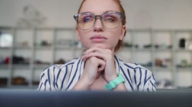 Düşünen kızıl saçlı, gözlüklü bir kadın laptopunu kullanarak dairenin içinde oturuyor.