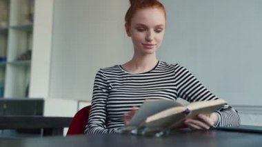 Güzel kızıl saçlı bir kadın evde oturmuş kitabı okuyor.