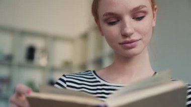 Çekici kızıl saçlı bir kadın evde oturmuş kitabı okuyor.