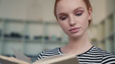 Odaklanmış kızıl saçlı bir kadın evde oturmuş kitabı okuyor.