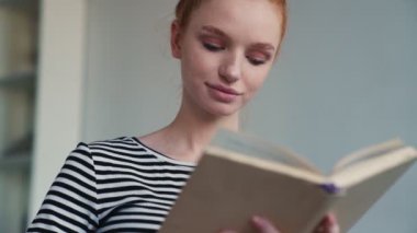 Gülümseyen kızıl saçlı bir kadın evde oturmuş kitabı okuyor.