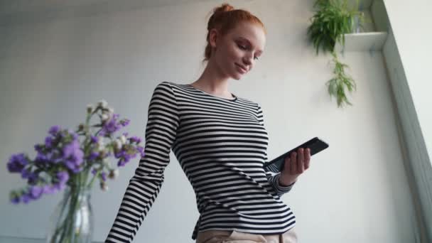 一个漂亮的红头发女人站在公寓里用她的智能手机 — 图库视频影像