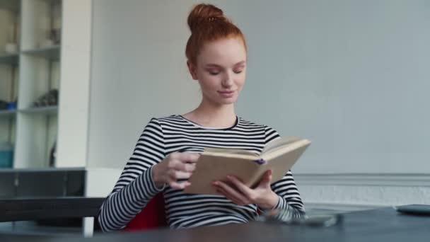 幸せな赤毛の女性は本を読んでいますアパートの中に座って — ストック動画