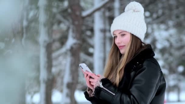冬の帽子をかぶったかなり素敵な若い女性が冬に外を歩く彼女のスマートフォンを使用しています — ストック動画