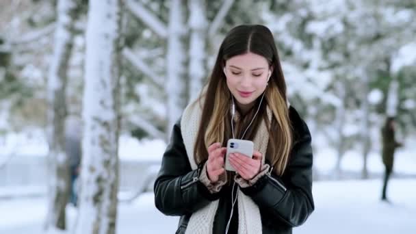 冬に外を歩いていると イヤフォンを持つ魅力的な若い女性がスマートフォンを使っています — ストック動画