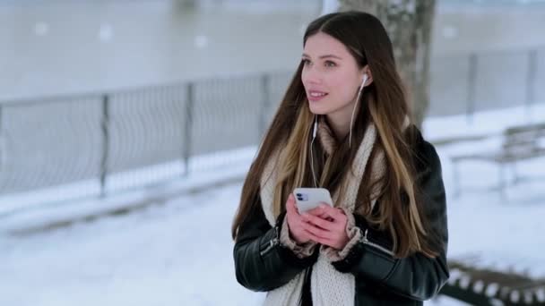 冬の公園の外を歩いていると イヤホンを持った幸せな若い女性がスマートフォンを使っています — ストック動画