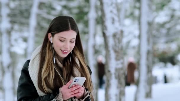 冬に外を歩いていると イヤホンを持った笑顔の若い女性がスマートフォンを使っています — ストック動画