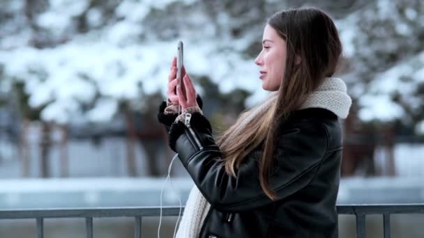 งสาวท ายร ปโดยใช สมาร ทโฟนของเธอขณะท นออกไปข างนอกในสวนสาธารณะในช วงฤด หนาว — วีดีโอสต็อก