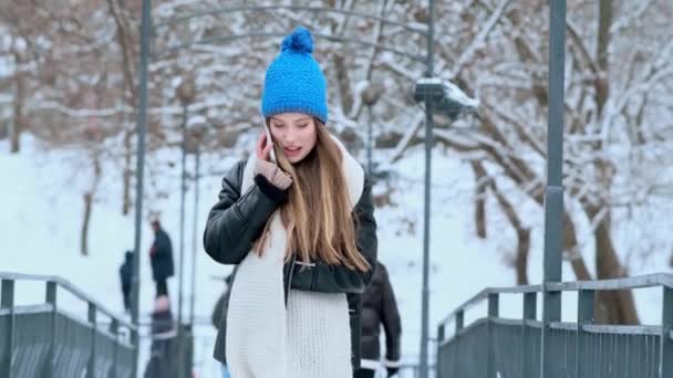 青い冬の帽子をかぶった笑顔の若い女性が冬の中央公園の外を歩くスマートフォンで話しています — ストック動画