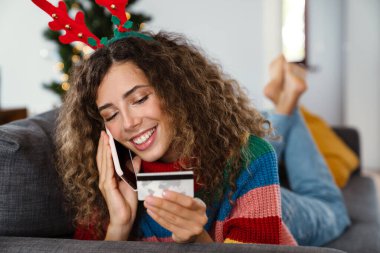 Mutlu kadın cep telefonuyla konuşuyor ve rahat bir odada kanepede yatarken elinde kredi kartı tutuyor.