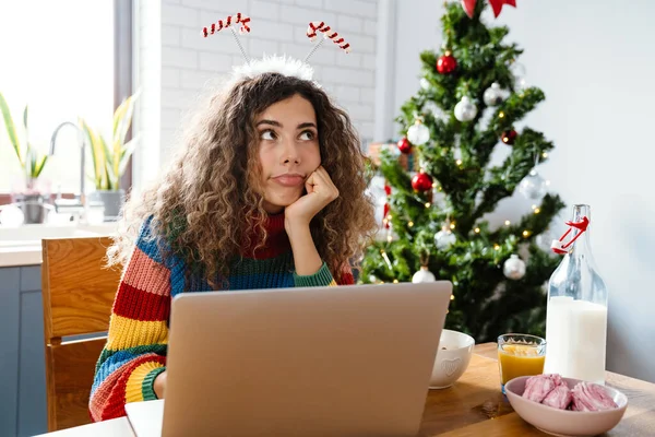 Thinking charming woman in toy horns using laptop while having breakfast in cozy kitchen