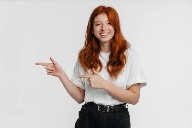 Ginger güzel mutlu kız gülümsüyor ve beyaz arka planda izole edilmiş parmakları işaret ediyor.