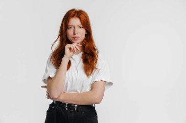 Kızıl güzel ciddi kız beyaz arka planda izole edilmiş kameraya bakıyor ve düşünüyor.