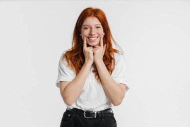 Ginger güzel neşeli kız gülümsüyor ve beyaz arka planda izole edilmiş yanaklarını işaret ediyor.