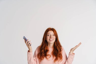 Ginger güzel neşeli kız cep telefonunu tutuyor ve beyaz arka planda izole bir şekilde elleri havaya kaldırıyor.